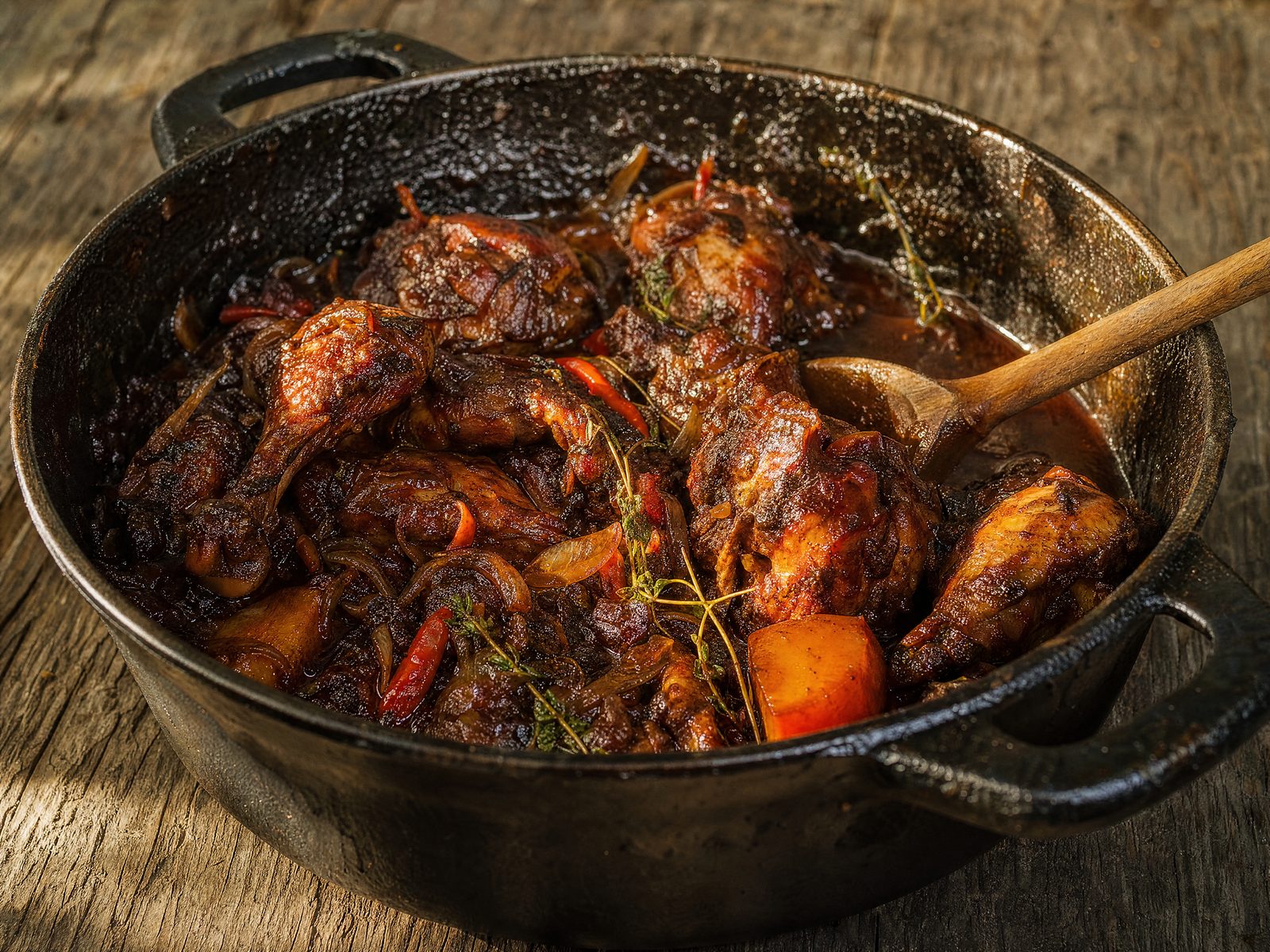 A black cast-iron pot of Trini brown stew chicken — dark, glossy mahogany-glazed chicken pieces in a rich gravy with sliced onions, strips of red pepper and fresh thyme sprigs, with a wooden spoon resting in the pot.