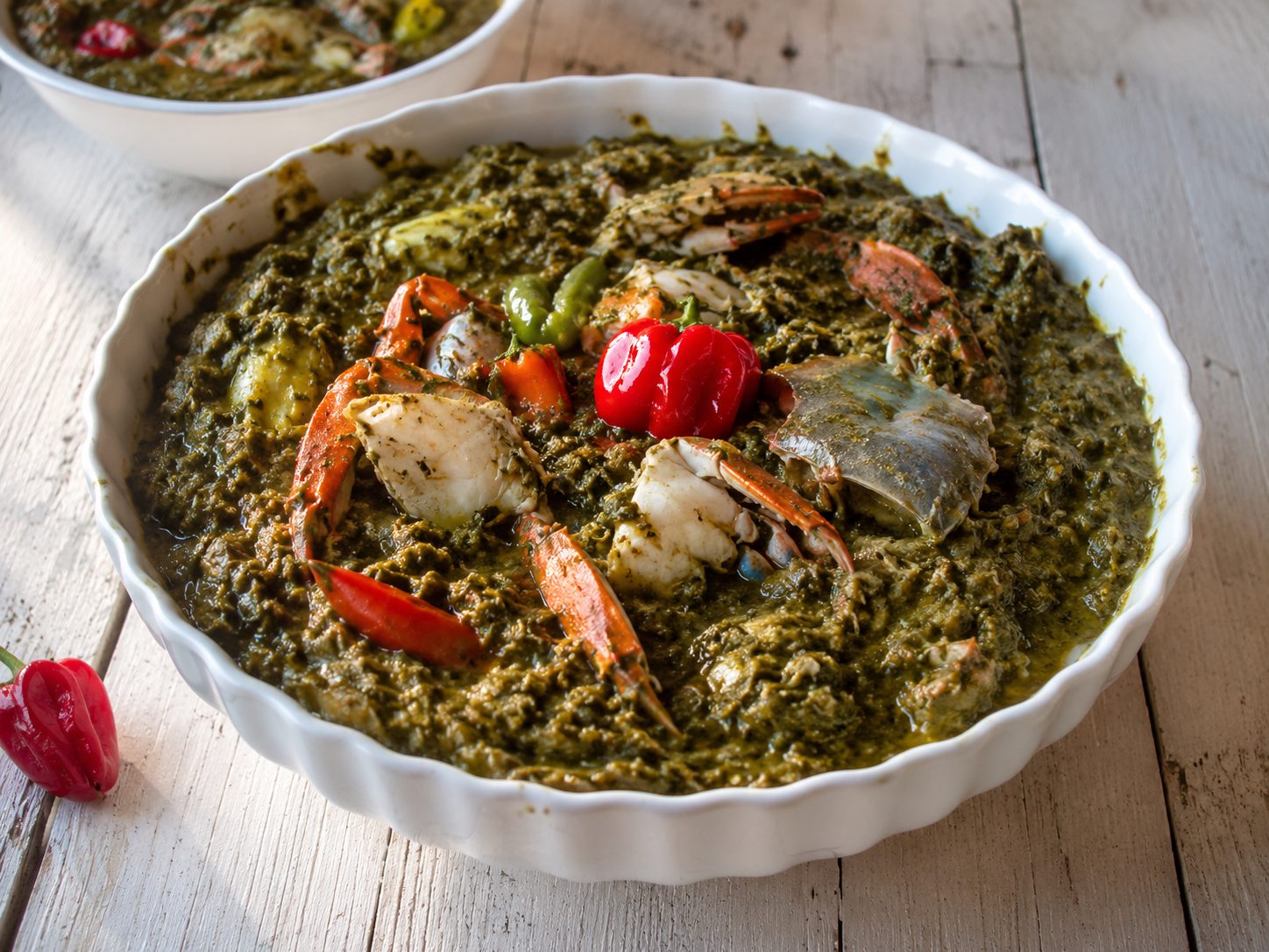 A white fluted bowl of thick, dark-green Trinidad callaloo topped with blue crab claws and shell, a whole red scotch bonnet pepper and a green pepper, on whitewashed wooden boards.