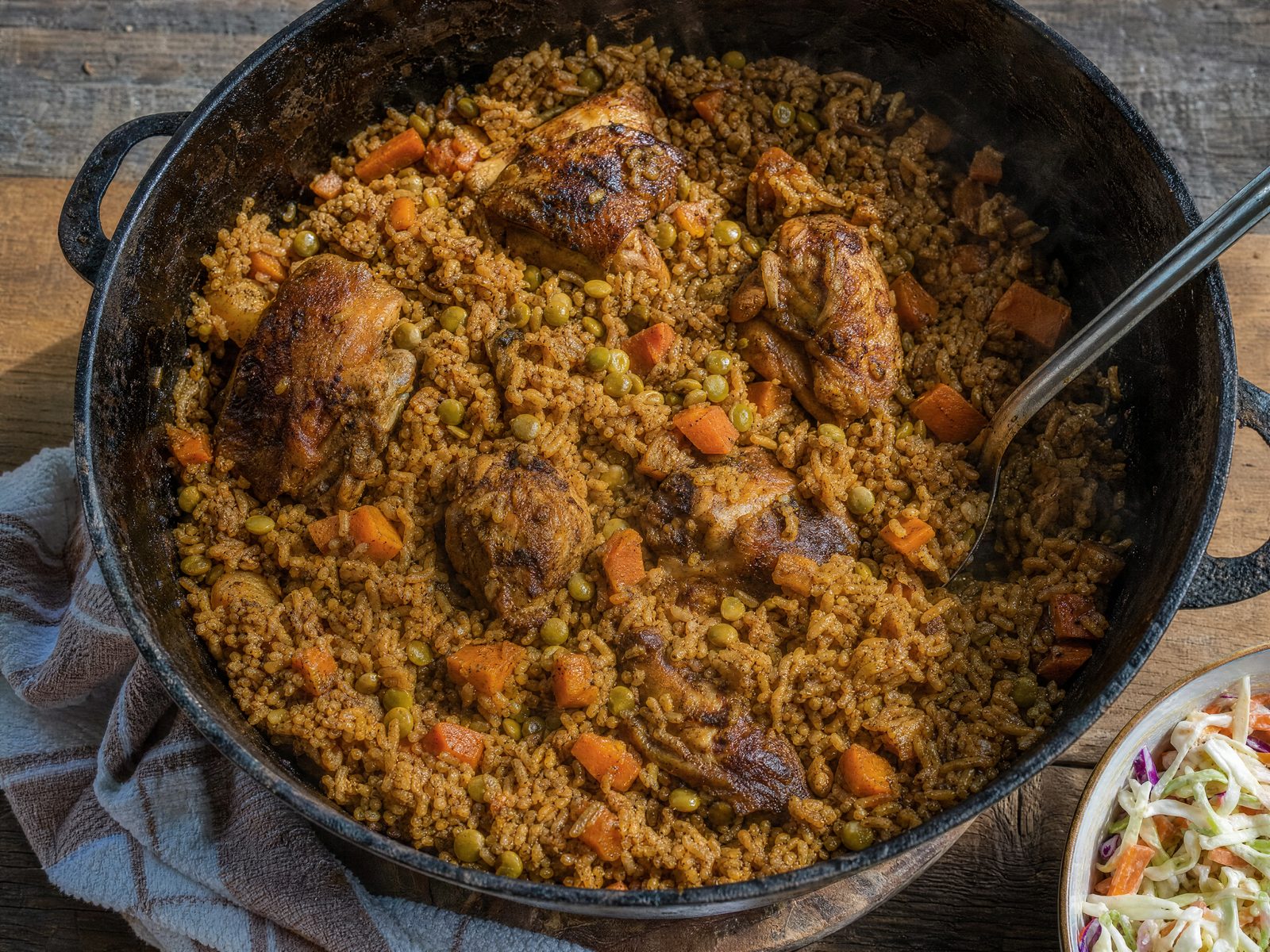 An overhead view of a black cast-iron pot of Trinidad chicken pelau — caramel-browned rice studded with green pigeon peas and chunks of carrot and pumpkin, with browned bone-in chicken pieces and a serving spoon, beside a bowl of coleslaw.