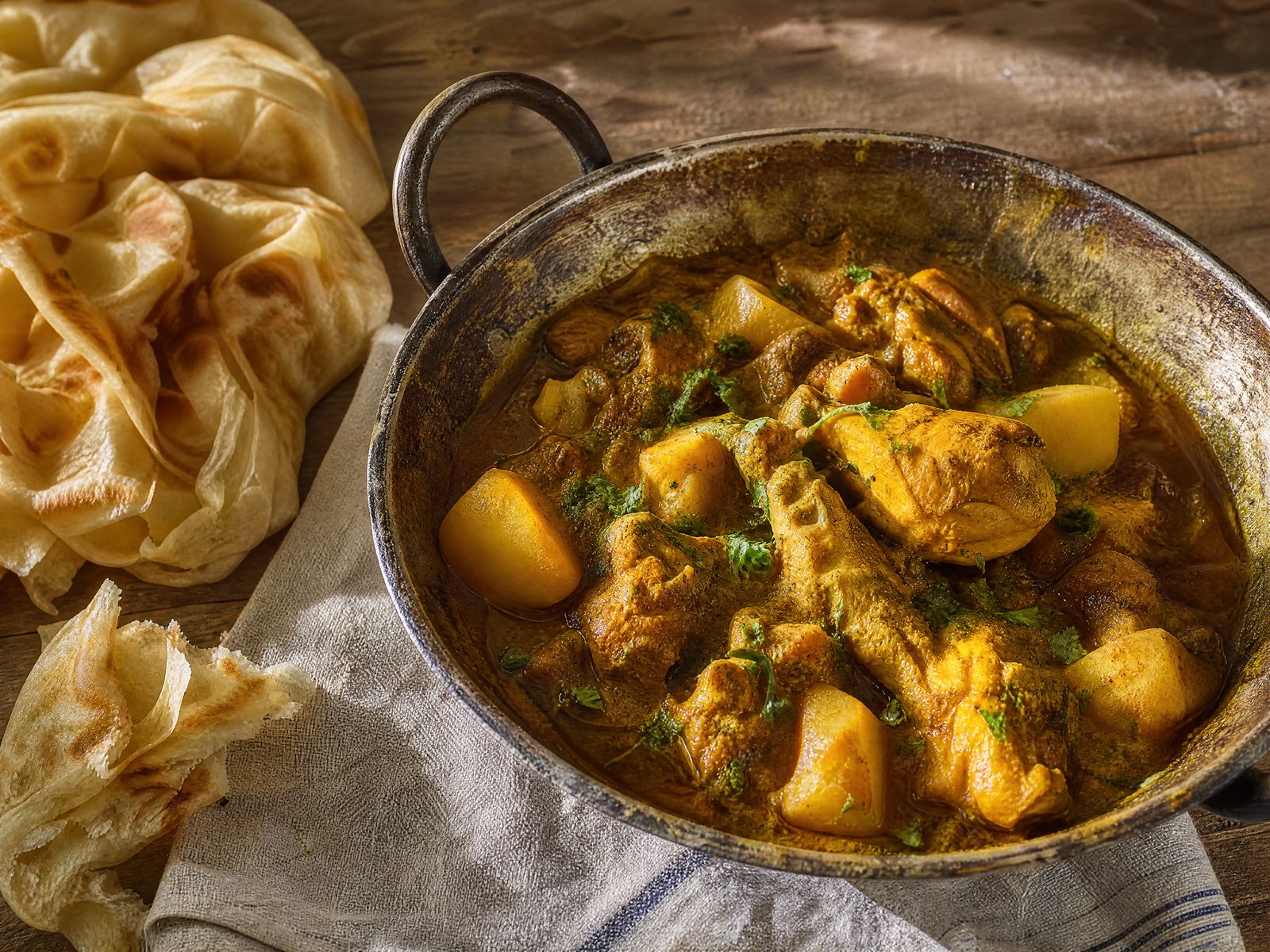 A brass karahi of golden Trini curry chicken with turmeric-yellow potatoes and scattered fresh cilantro, served alongside torn pieces of flaky buss-up-shut roti on a striped cloth.
