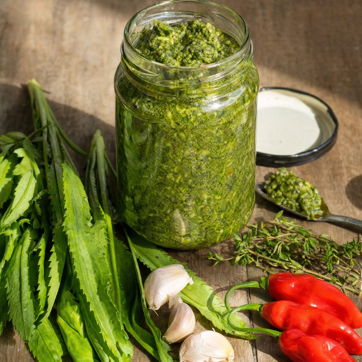 An open glass jar packed with coarse, bright-green Trinidad green seasoning, surrounded by fresh chadon beni leaves, garlic cloves, thyme sprigs, scallions and red pimento peppers on a weathered wooden table.